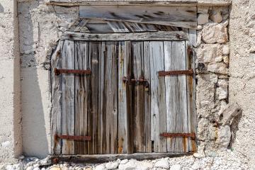 A close-up view of a weathered, grey wooden double door set into a rough, white-washed stone wall, with a padlock securing the center and rusted metal hinges visible.