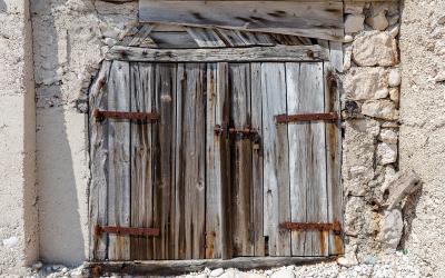 A close-up view of a weathered, grey wooden double door set into a rough, white-washed stone wall, with a padlock securing the center and rusted metal hinges visible.