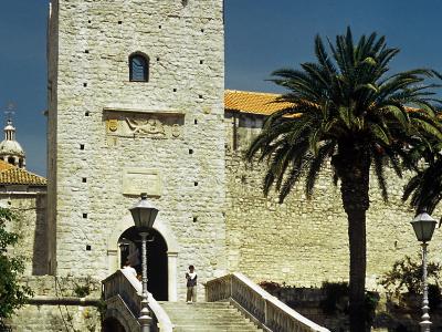 A tall, rectangular stone tower with crenellations, an arched entrance accessed by a wide stone staircase, a palm tree to the right, and a flagpole on top, set against a clear blue sky.