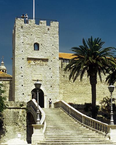 A tall, rectangular stone tower with crenellations, an arched entrance accessed by a wide stone staircase, a palm tree to the right, and a flagpole on top, set against a clear blue sky.