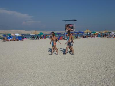 A wide, pebbly beach scene with numerous colorful umbrellas, beachgoers, and a lifeguard tower featuring a large advertisement in the background under a clear blue sky.