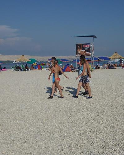 A wide, pebbly beach scene with numerous colorful umbrellas, beachgoers, and a lifeguard tower featuring a large advertisement in the background under a clear blue sky.
