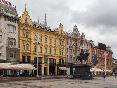 The image shows a wide pedestrian square featuring a prominent equestrian statue on a stone pedestal, surrounded by ornate 19th-century European-style buildings, including a distinctive yellow building with green domes and a shop with a "Jana" awn