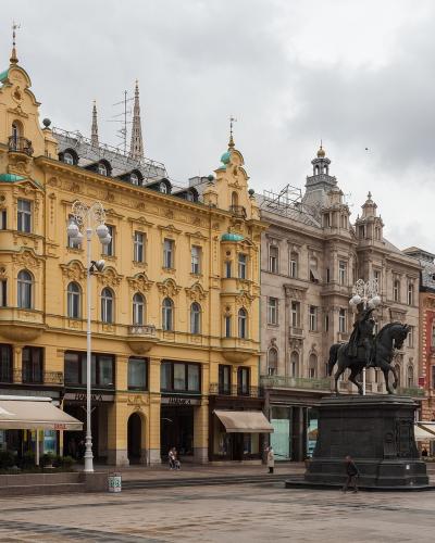 The image shows a wide pedestrian square featuring a prominent equestrian statue on a stone pedestal, surrounded by ornate 19th-century European-style buildings, including a distinctive yellow building with green domes and a shop with a "Jana" awn
