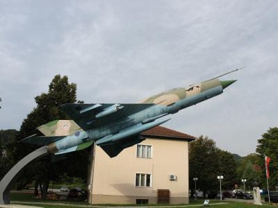 A MiG-21 fighter jet mounted on a curved pedestal as a monument, situated in a grassy public area next to a beige building, with trees, parked cars, and a Croatian flag visible in the background.