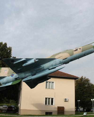 A MiG-21 fighter jet mounted on a curved pedestal as a monument, situated in a grassy public area next to a beige building, with trees, parked cars, and a Croatian flag visible in the background.
