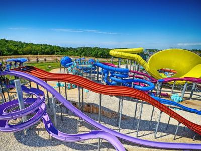 A high-angle view of a large outdoor water park featuring multiple colorful slides (purple, red, blue, yellow) on a gravel base with green hills in the background under a clear blue sky.