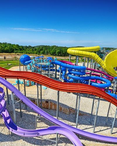 A high-angle view of a large outdoor water park featuring multiple colorful slides (purple, red, blue, yellow) on a gravel base with green hills in the background under a clear blue sky.