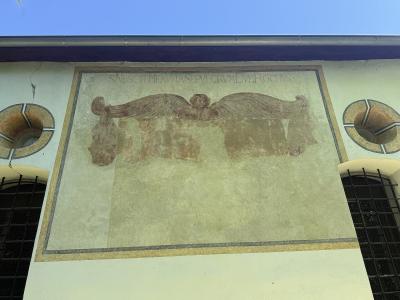 The image shows a weathered mural on an exterior wall featuring a winged cherub holding a banner with the Latin inscription "S.NESCIT ME AVT ASE PVLGRVM DVM ROCHVS", flanked by two circular decorative elements and barred windows below.