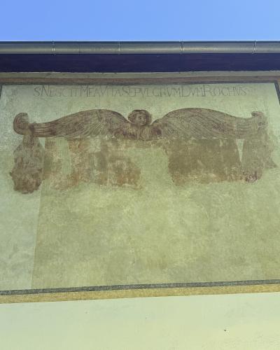 The image shows a weathered mural on an exterior wall featuring a winged cherub holding a banner with the Latin inscription "S.NESCIT ME AVT ASE PVLGRVM DVM ROCHVS", flanked by two circular decorative elements and barred windows below.